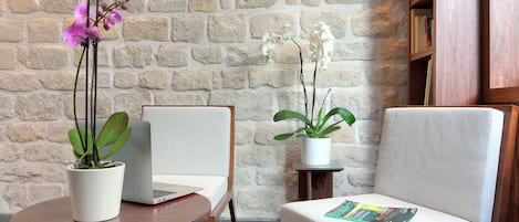 Una habitación moderna con una pared de piedra, una mesa redonda de madera con un ordenador portátil y una orquídea en maceta, una silla blanca con cojín y una revista, y otra planta en maceta.