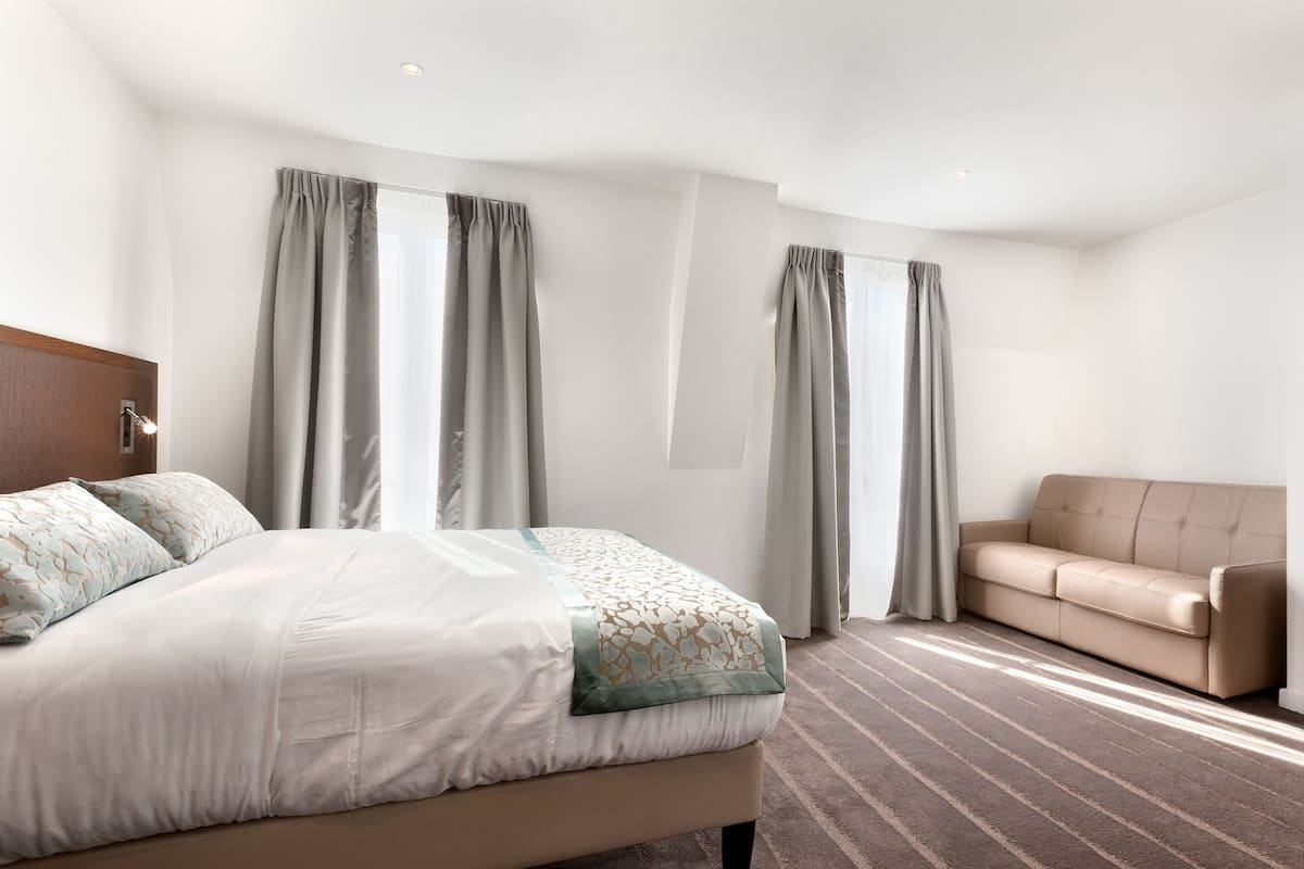 A hotel room with a bed, a sofa, and large windows with curtains.
