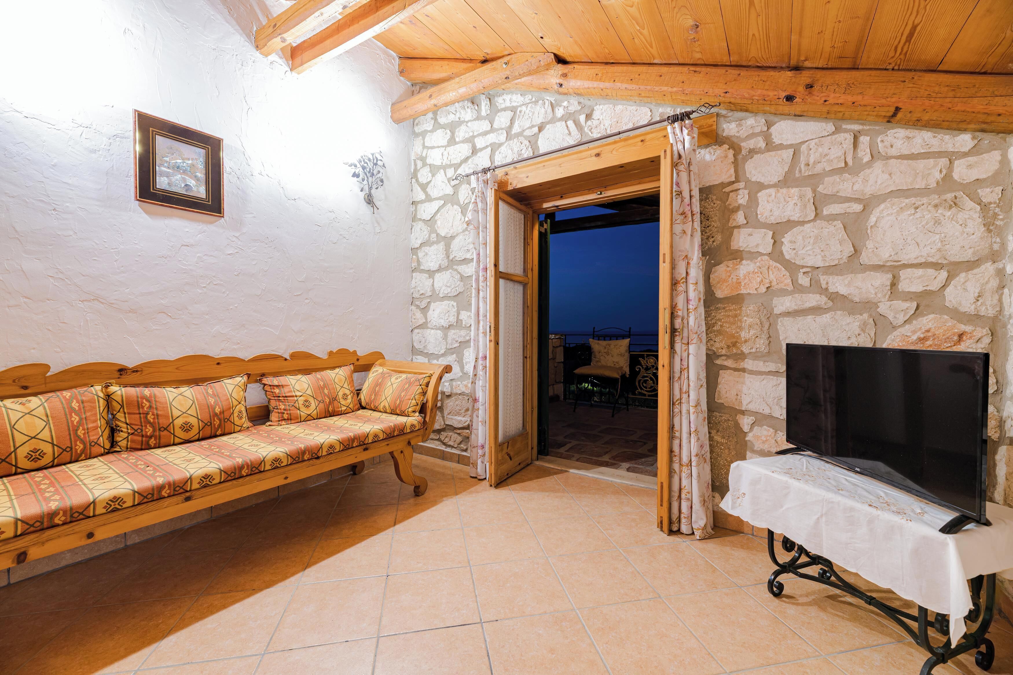 A stone-walled room with a wooden ceiling, a sofa, a TV on a stand, and an open doorway leading outside.
