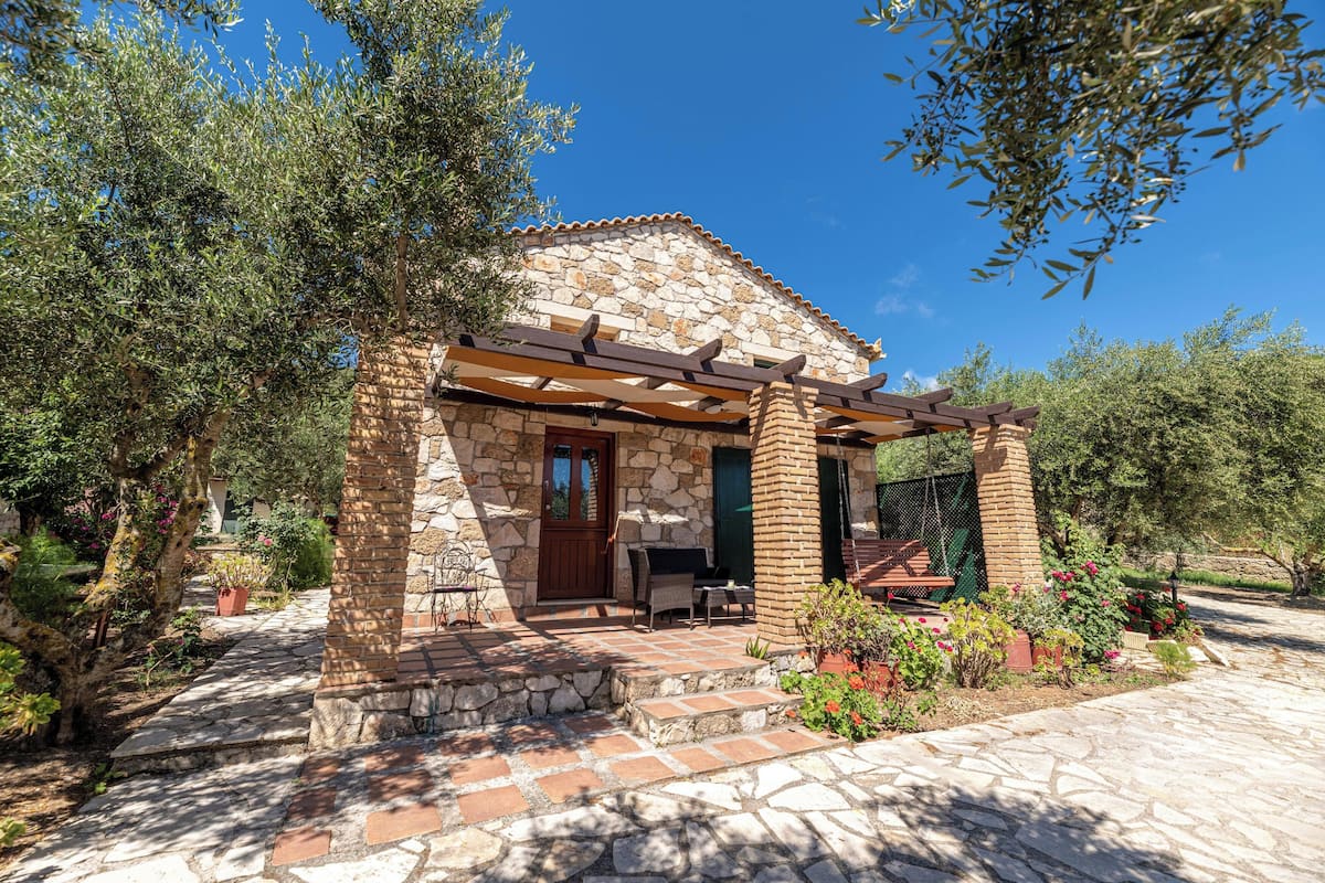 A stone house with a wooden pergola, a stone pathway, and surrounding greenery.