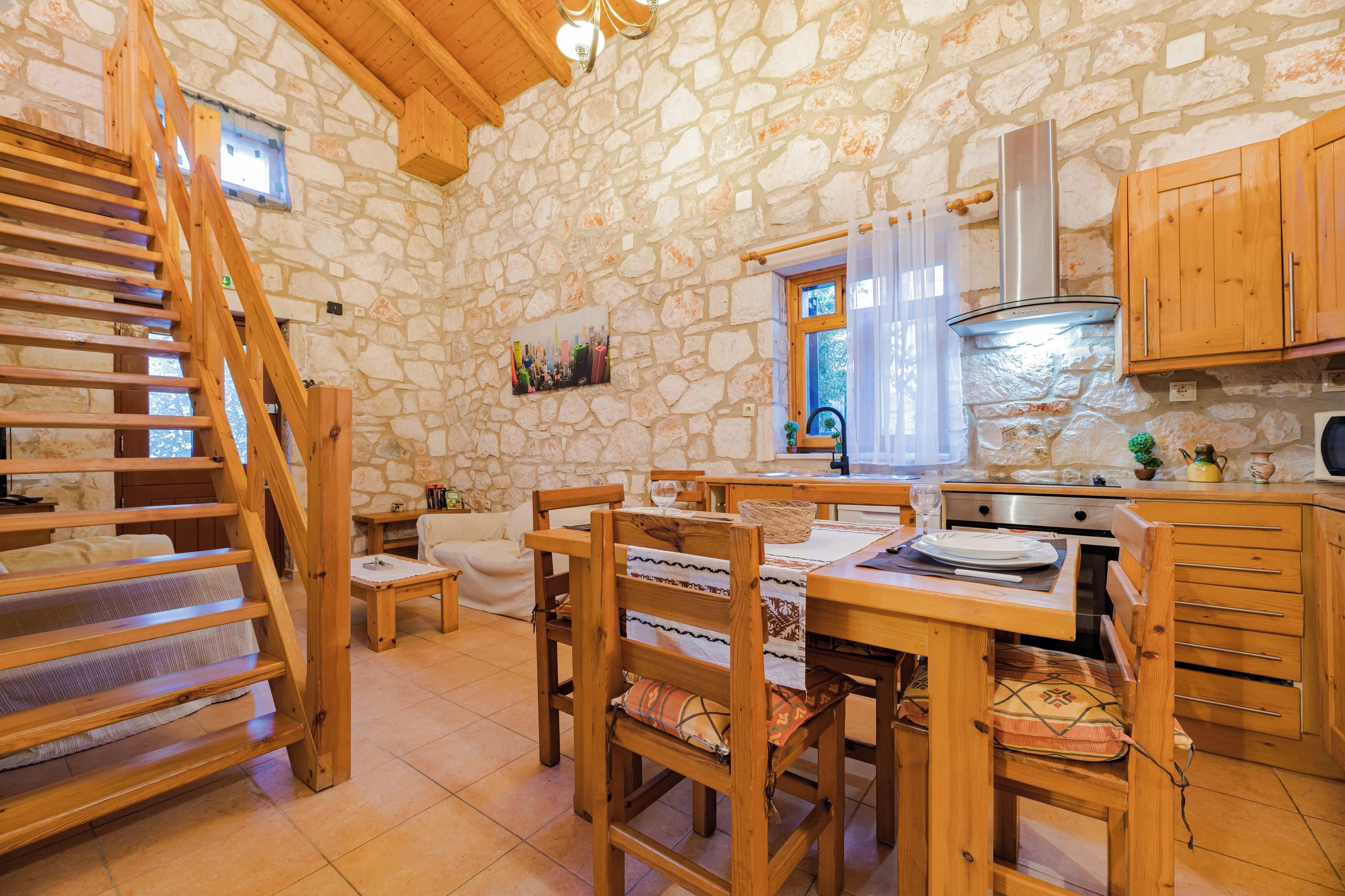 A stone-walled kitchen with wooden furniture, a staircase, and a window with curtains.