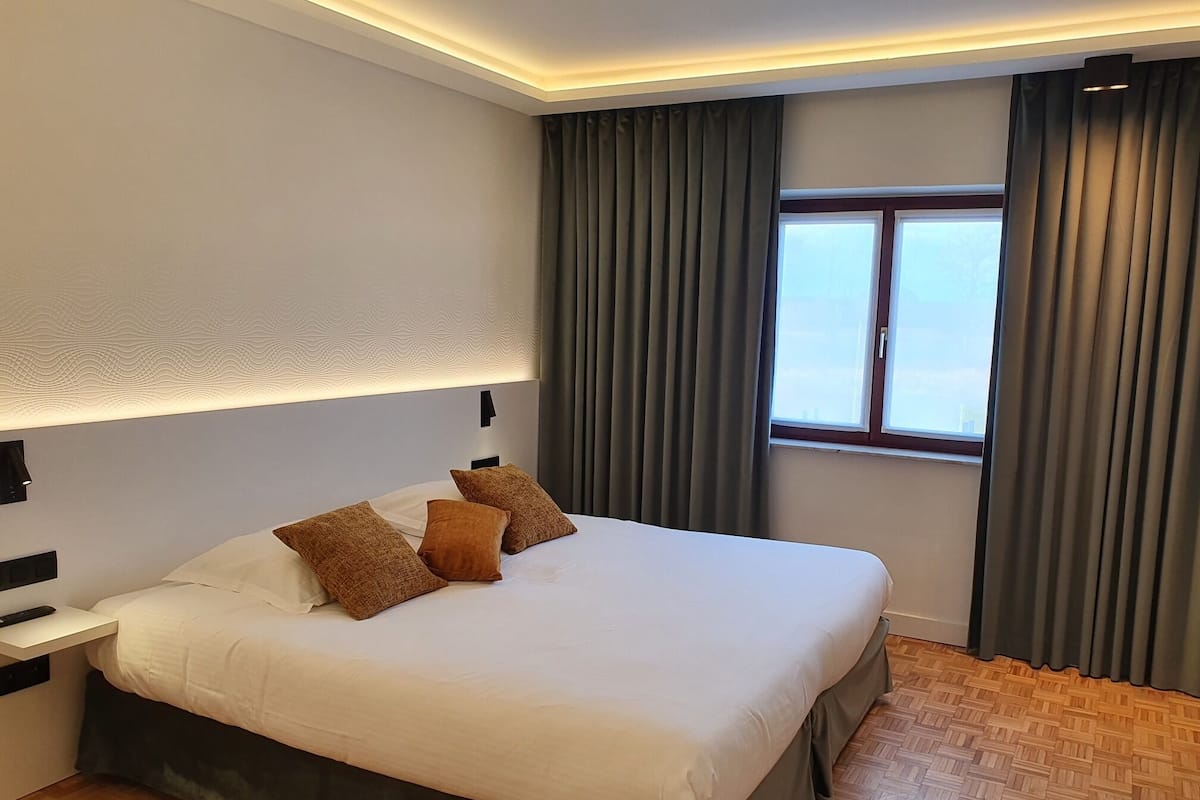 A neatly made bed with white and brown pillows, a bedside table with a remote control, and a window with curtains.