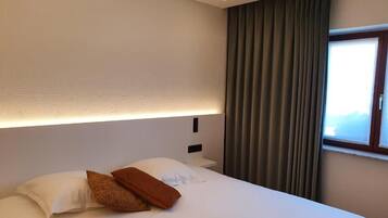 Quarto de hotel com cama bem arrumada, cabeceira com iluminação embutida, janela com cortinas e vista para o exterior.