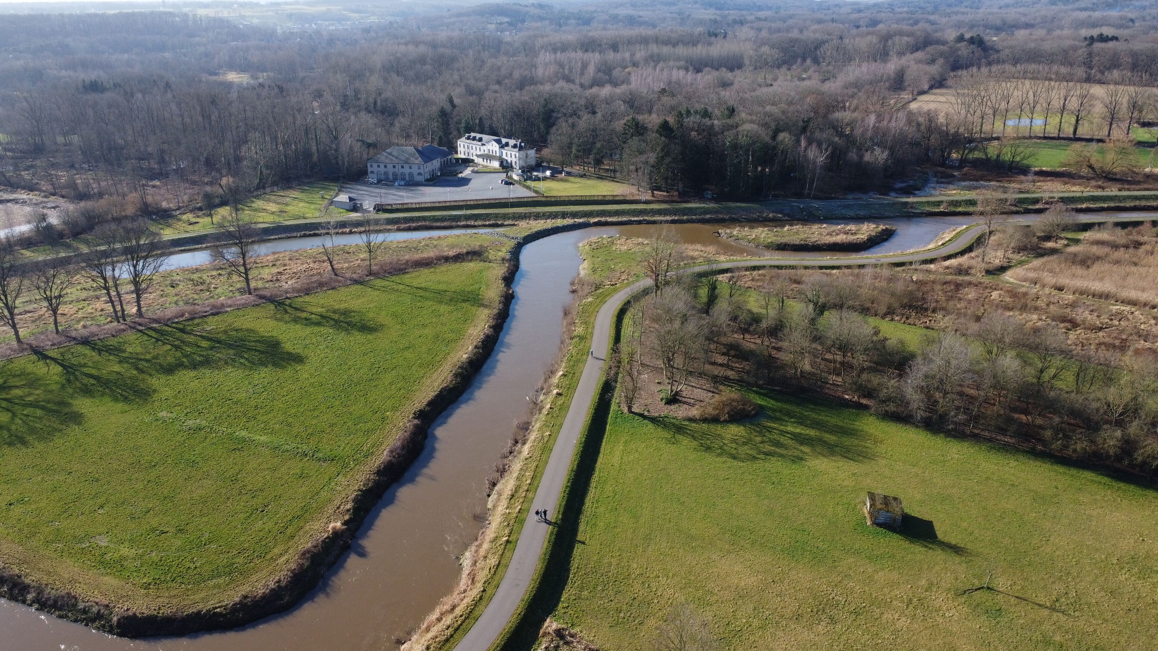 Une rivière serpente à travers un paysage rural, bordée d’une grande propriété et d’un chemin pavé.
