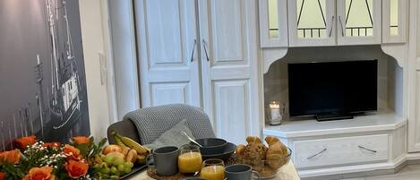 A modern living room with a dining area, featuring a large table set for breakfast, a flat-screen TV, and a decorative wall piece.