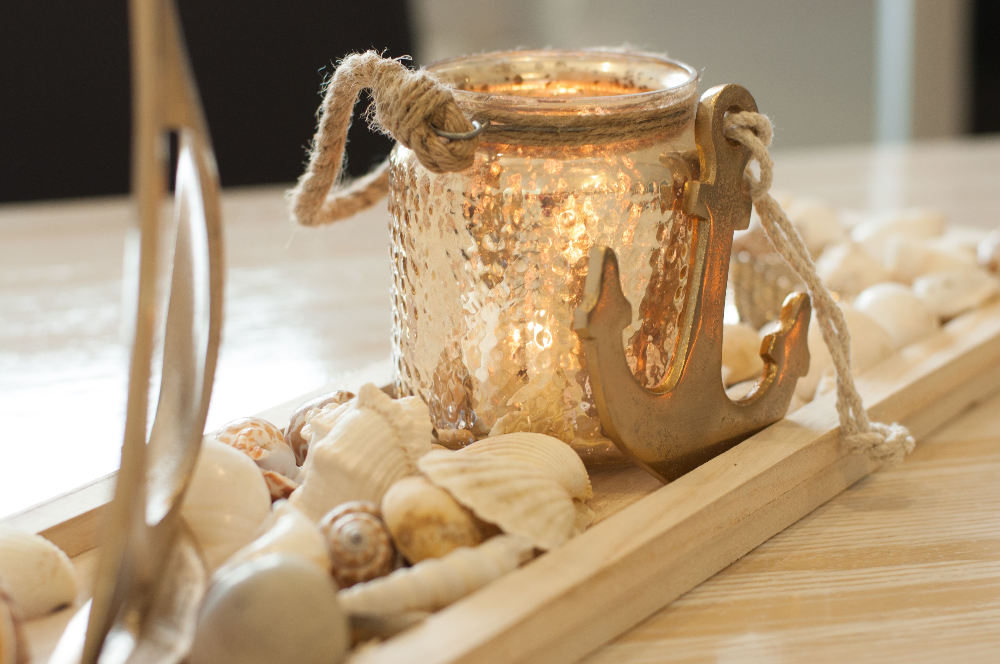 A decorative glass candle holder with a rope handle, surrounded by seashells on a wooden tray.