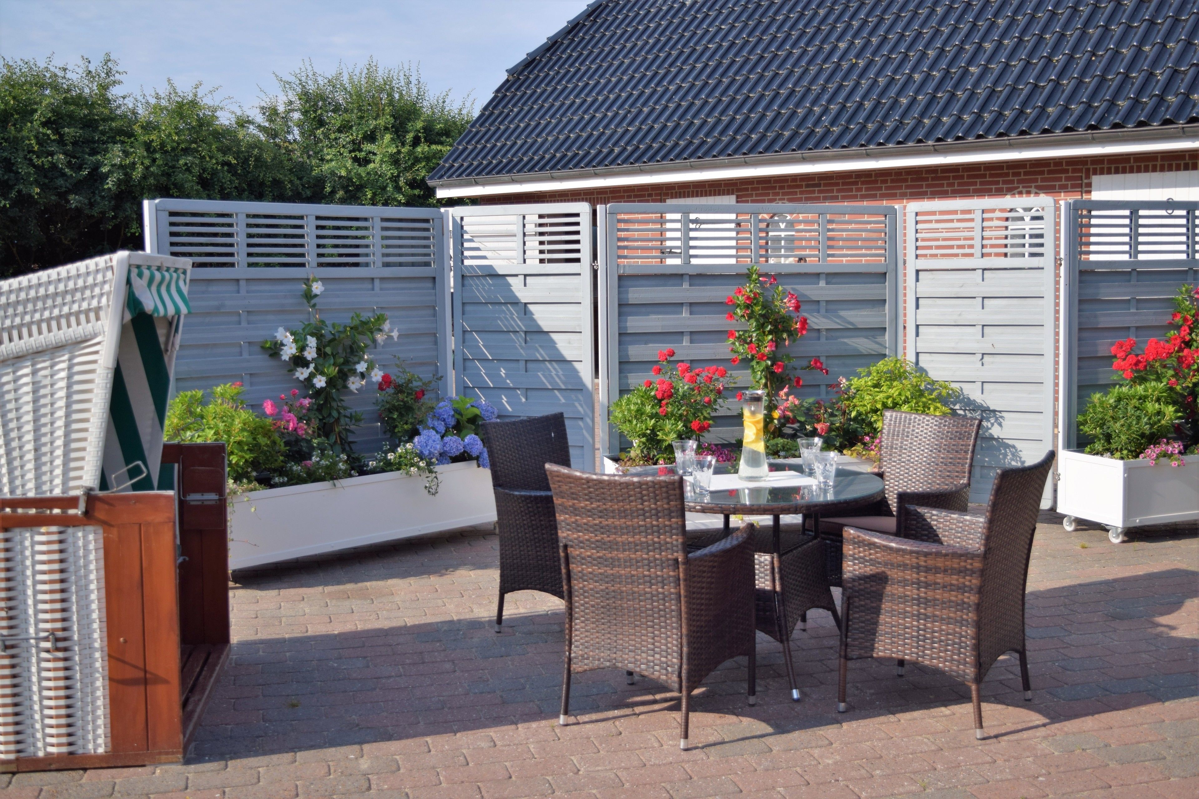 Une terrasse extérieure avec des chaises et une table en osier, entourée d’une clôture blanche et de plantes en pot.