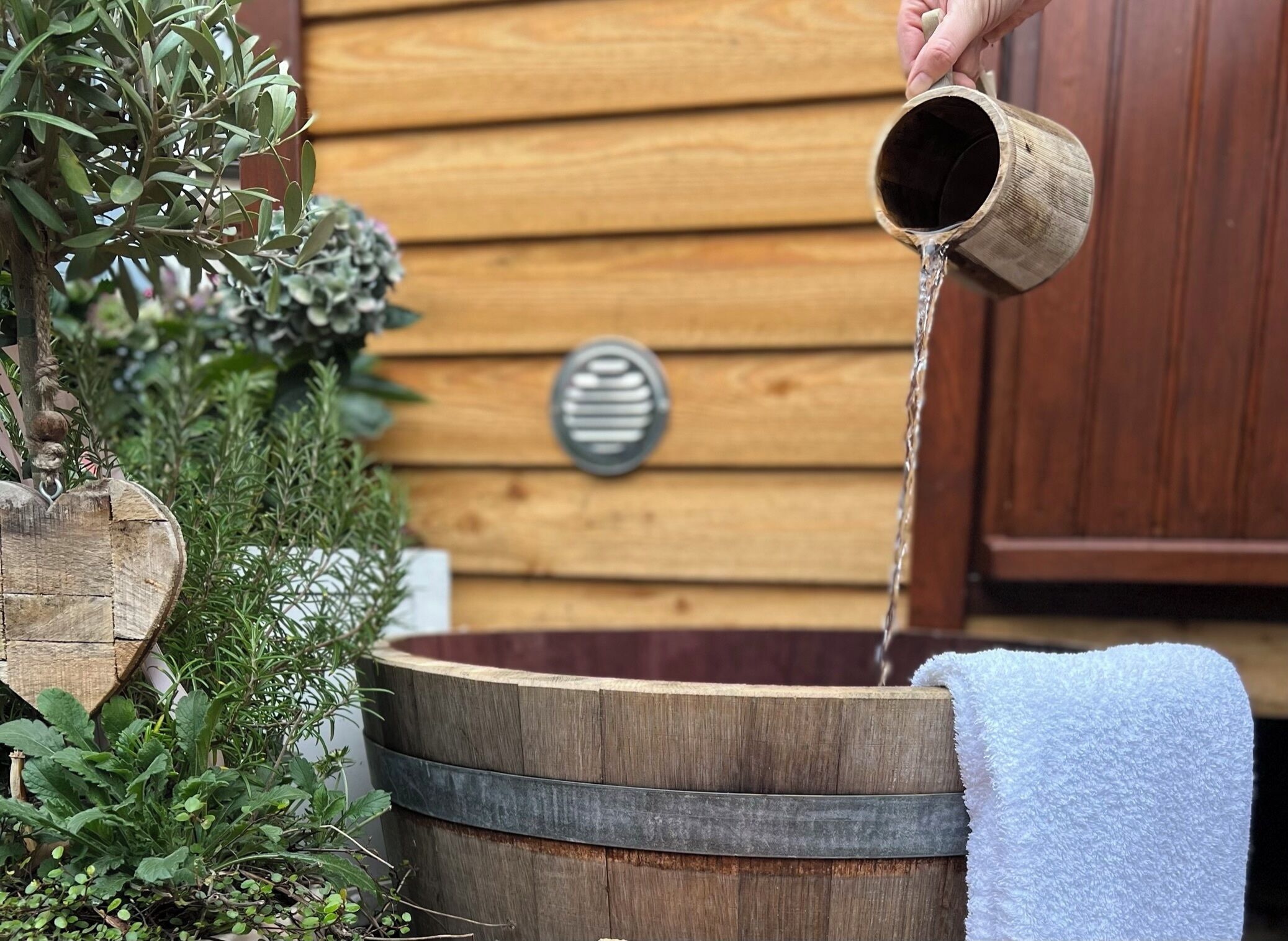 A wooden barrel with water flowing into it, placed outdoors with a wooden wall and plants in the background.