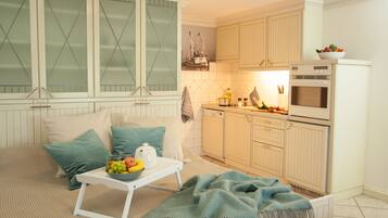 A cozy room with a bed, a small table with a bowl of fruit, a kettle, and a blanket. There is a kitchenette with a microwave, oven, and a bowl of fruit on the counter.
