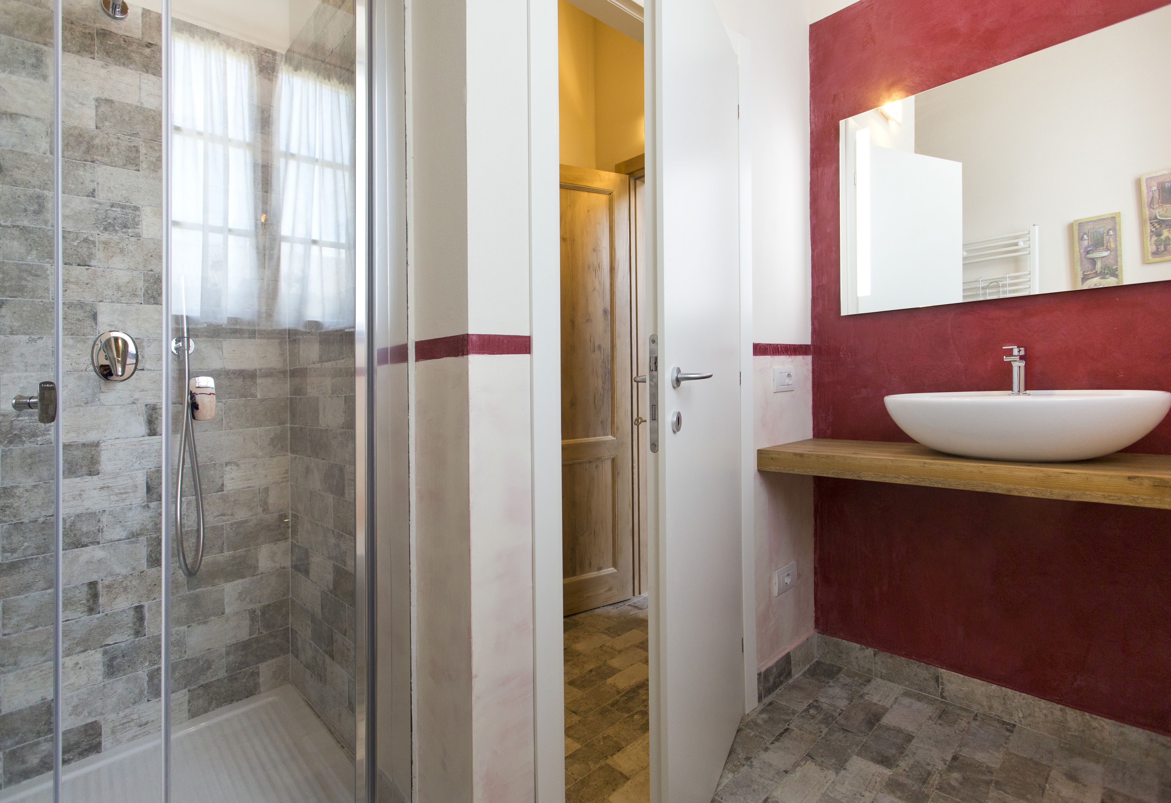 Un baño con cabina de ducha de vidrio, una puerta de madera, un lavabo blanco y una pared roja de acento, y un espejo sobre el lavabo.