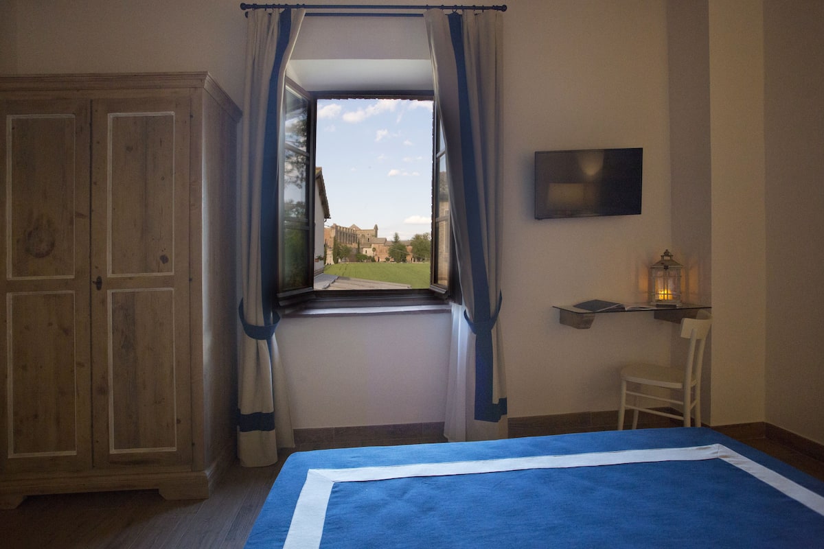 A bedroom with a large window, wooden wardrobe, bedside table with a lamp, and a TV on the wall.
