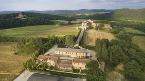 A large, historic building complex with multiple structures, surrounded by lush green fields and trees, under a partly cloudy sky.