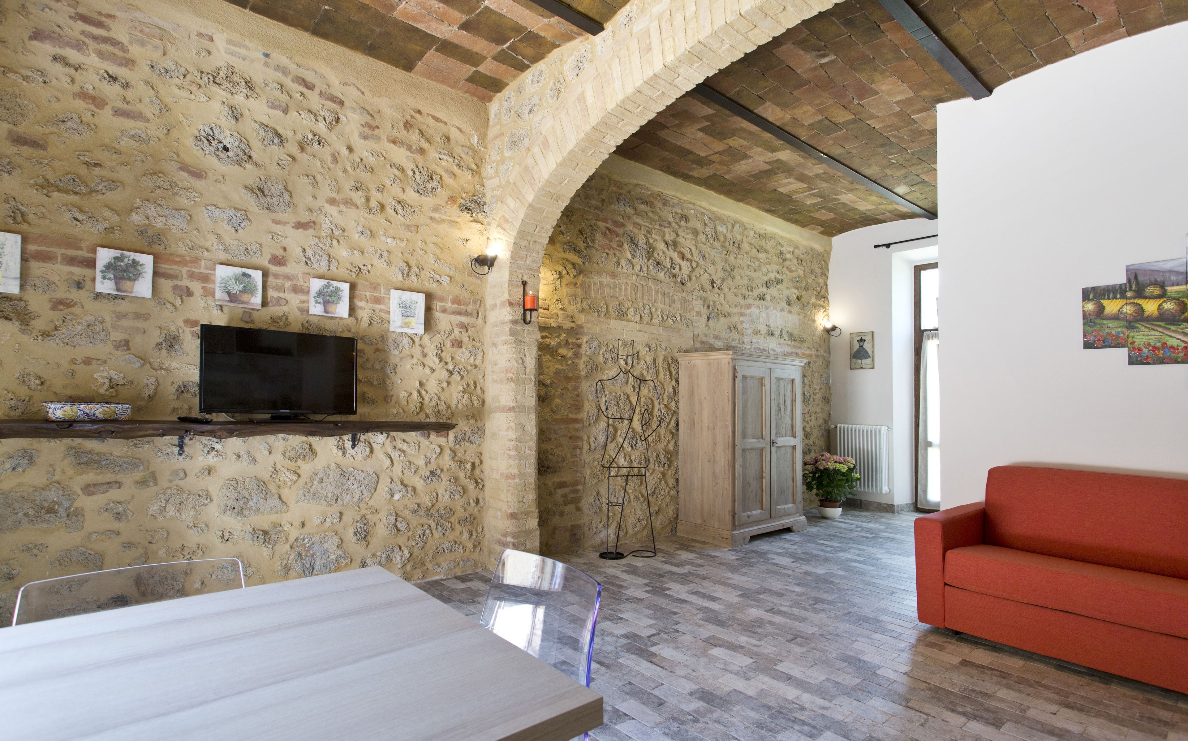 A rustic interior with exposed brick walls, a wooden table, a red sofa, and a television mounted on the wall.