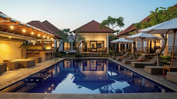 A swimming pool with lounge chairs and umbrellas, a covered patio area, and a building with a tiled roof.