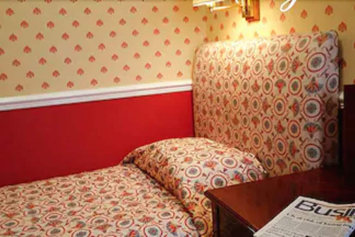 Een knus kamertje met een enkel bed, een hoofdbord met een patroon en een tafel met een krant.