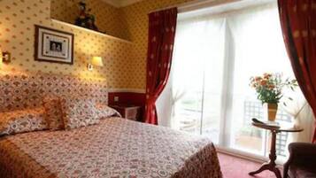 Een hotelkamer met een bed met een patroon, een nachtkastje, een bijzettafeltje met een lamp, een ingelijst schilderij aan de muur en een raam met gordijnen.