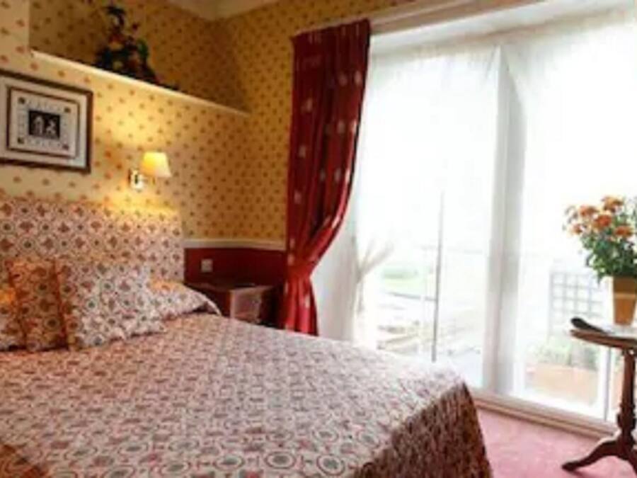 Een hotelkamer met een bed met een patroon, een nachtkastje, een bijzettafeltje met een lamp, een ingelijst schilderij aan de muur en een raam met gordijnen.