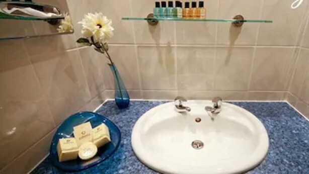 Une salle de bain avec un lavabo blanc, un plan de toilette bleu et une étagère en verre au-dessus du lavabo où sont rangés les articles de toilette.