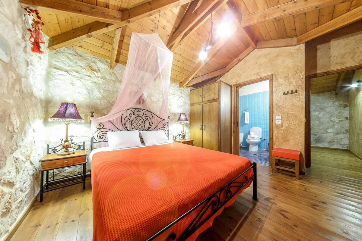A bedroom with a wooden ceiling, stone walls, a bed with an orange bedspread, a nightstand with a lamp, and a wooden bench.