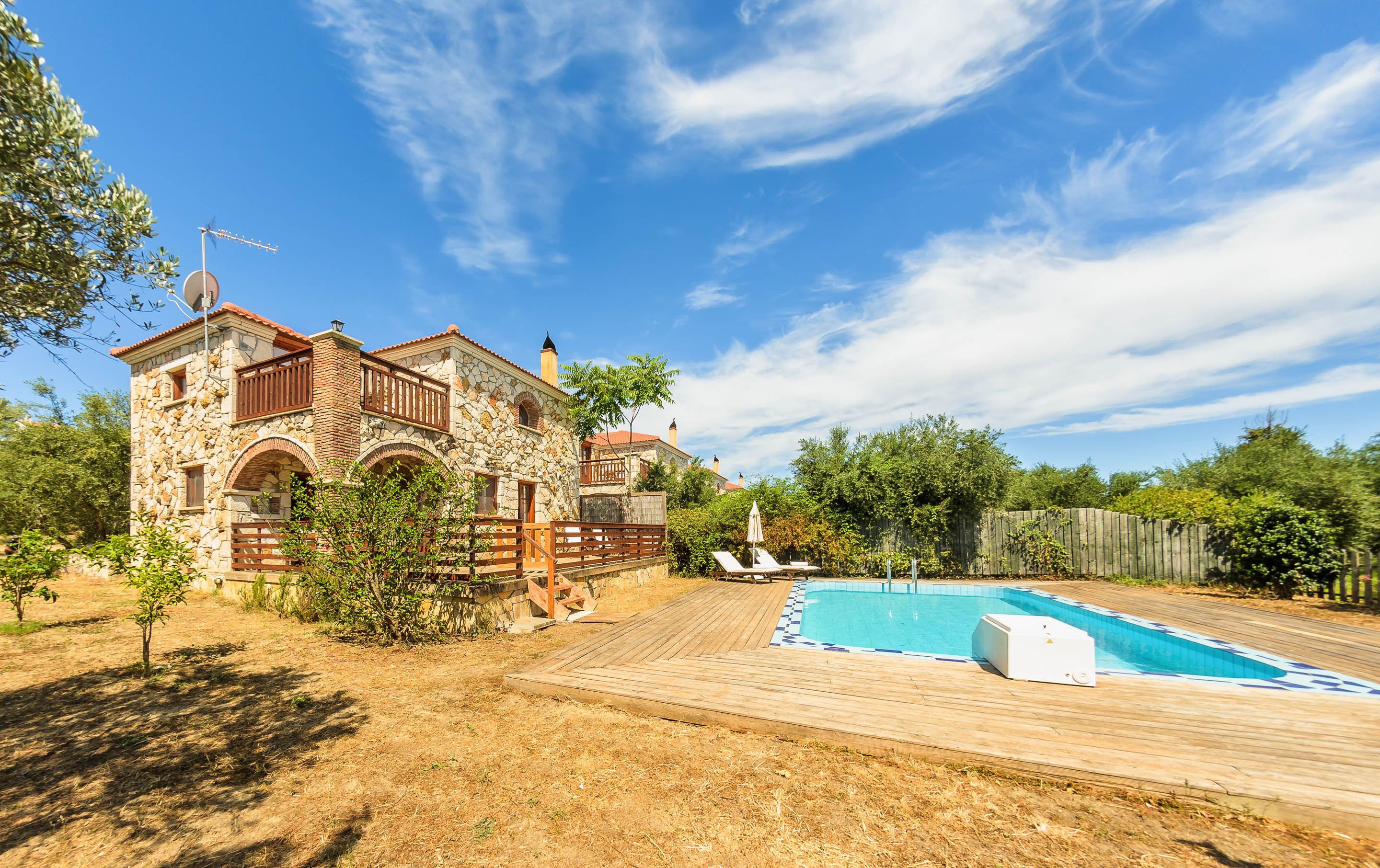 Une maison en pierre avec une piscine, entourée d’une terrasse en bois et d’un jardin clos.
