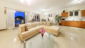 A spacious living room with a large beige sectional sofa, a glass coffee table with a vase of flowers, and a view of the sea through the open sliding doors. - Parys Villas (Zakynthos)