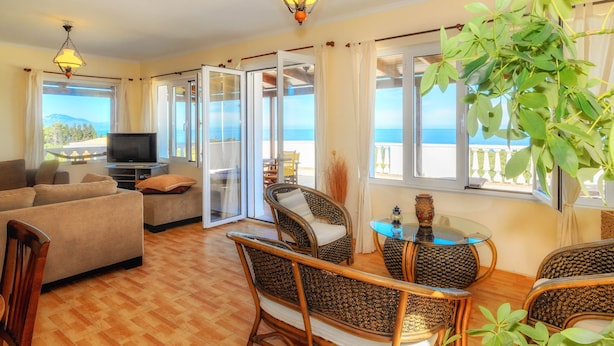 A spacious living room with a sofa, chairs, a TV, and a view of the sea through large windows. - Parys Villas (Zakynthos)