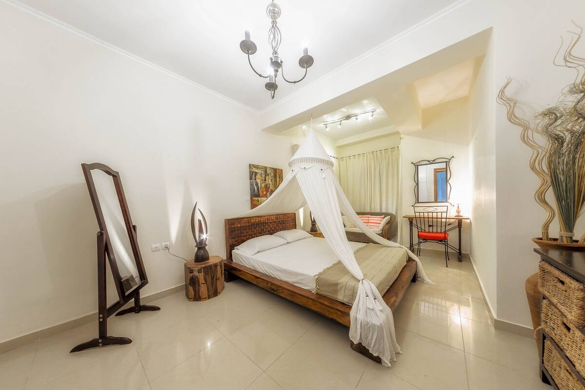 A bedroom with a four-poster bed, a standing mirror, a bedside table with a lamp, and a dresser.