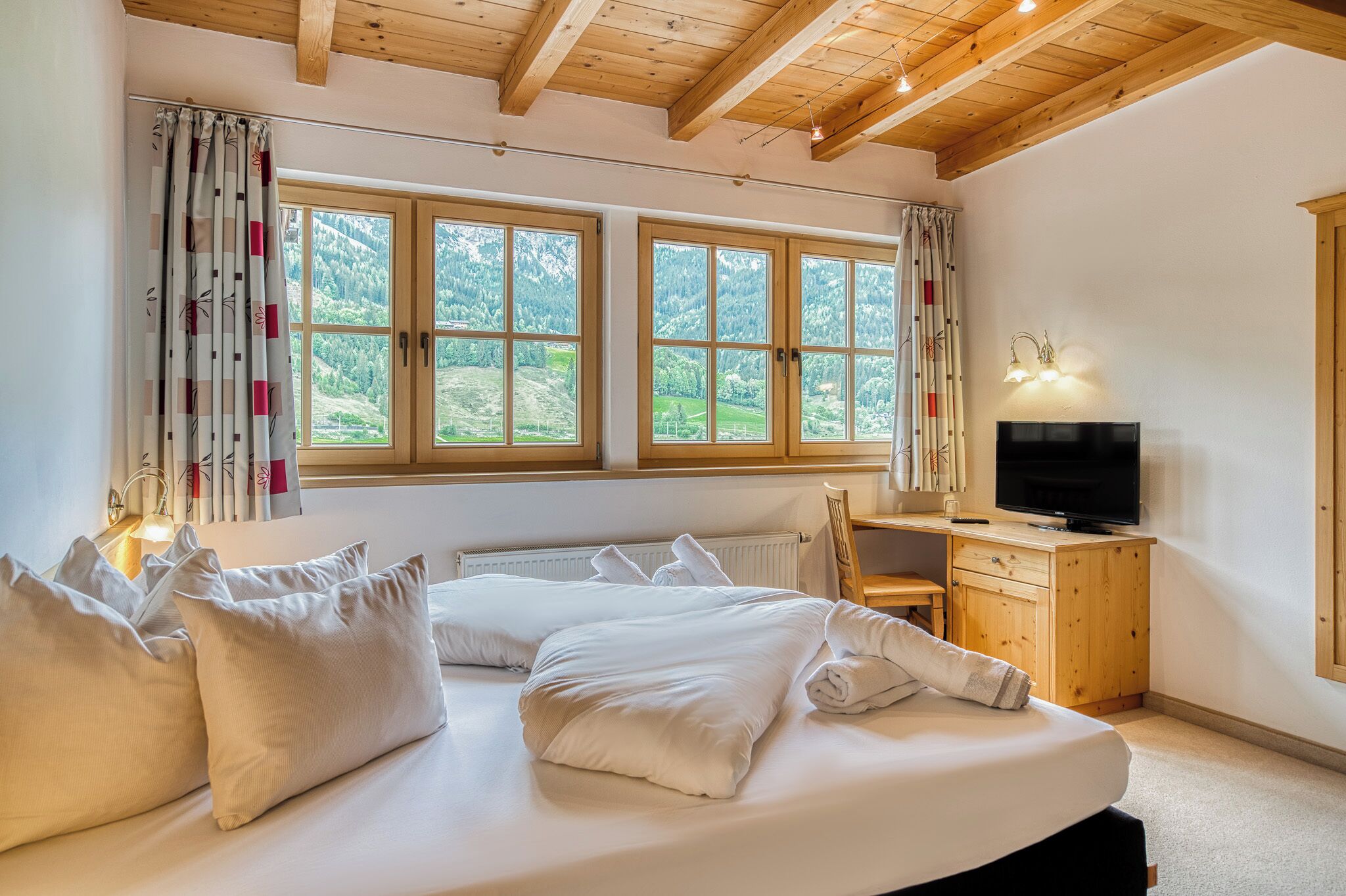 Une chambre à coucher avec un lit, un bureau, une télévision et de grandes fenêtres avec des rideaux.