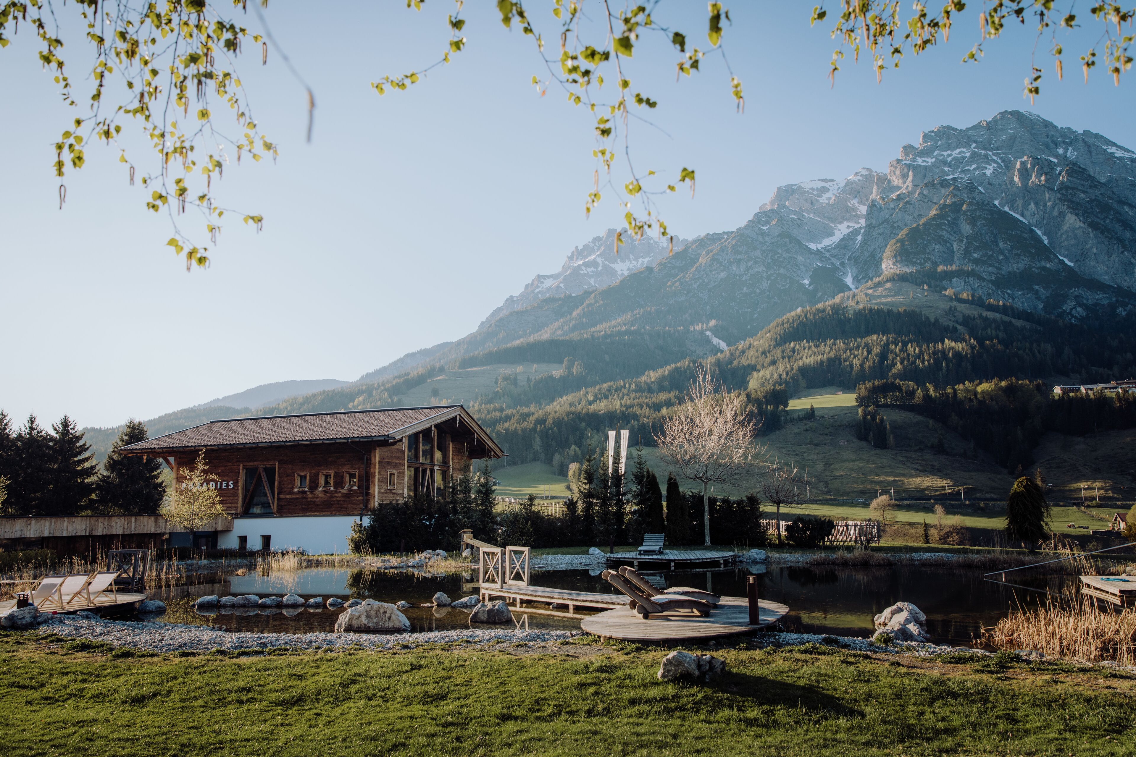 A rustic lodge with a wooden exterior, a pond, and a mountain backdrop.