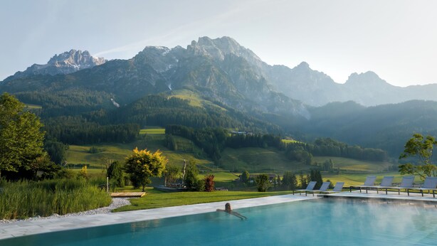 Une piscine avec une personne dans l’eau, entourée d’un espace paysager agrémenté d’arbres et offrant une vue sur des montagnes en arrière-plan.