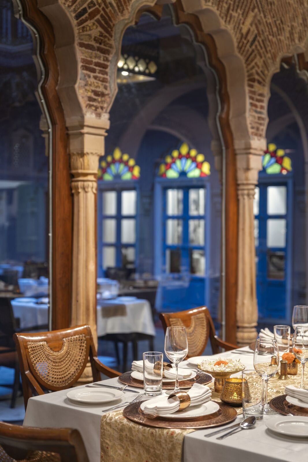 A restaurant with a traditional interior, featuring arched brick walls and stained glass windows. Tables are set with white tablecloths, gold-rimmed plates, and clear glassware.
