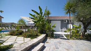 Une cour dallée de pierres, avec des marches menant à une porte blanche, entourée de plantes tropicales et d’une piscine.
