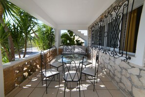 A sunlit patio with a stone wall, a glass-top table surrounded by chairs, and a view of the outdoors. - Mirsini Studios & Apartments (Zakynthos)