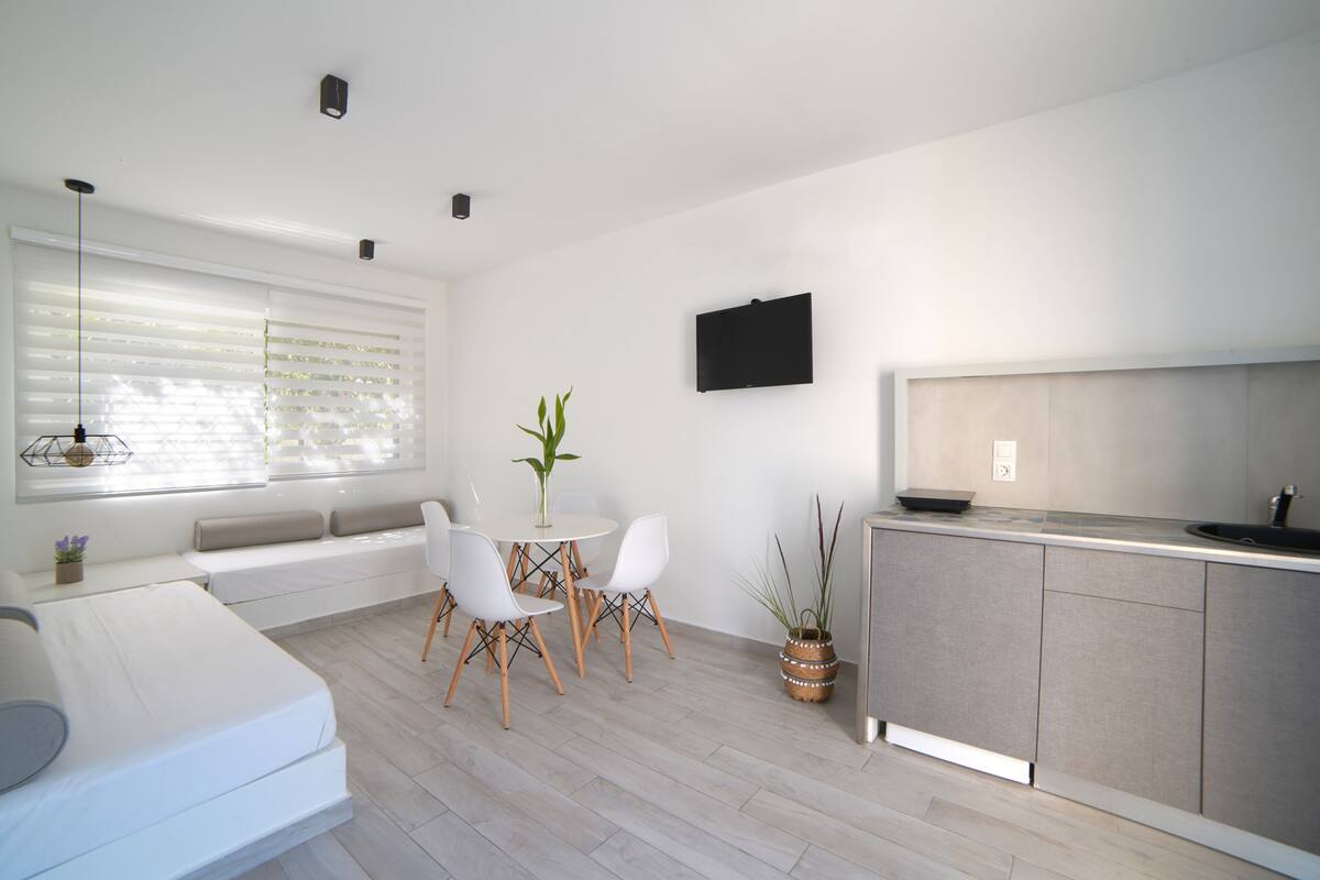 A modern kitchen with a dining area, white walls, and a large window with blinds.