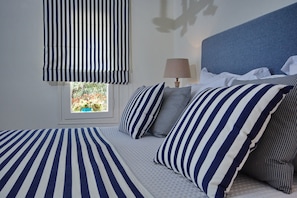 A bedroom with a striped bedspread, matching pillows, and a window with a view of greenery.