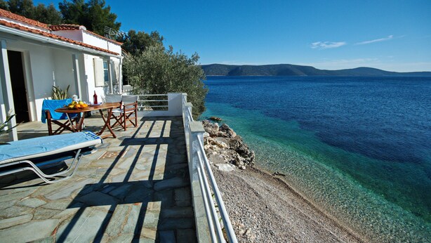 Una casa costera con un patio que da a un mar azul y transparente, una mesa con frutas y una silla.