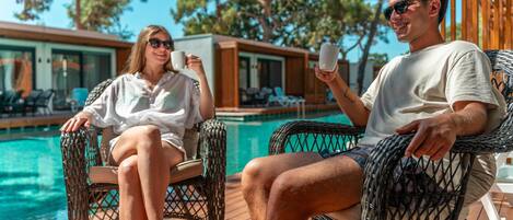 To personer sidder ved en poolliggestol, med en pool og træterrasse i baggrunden.