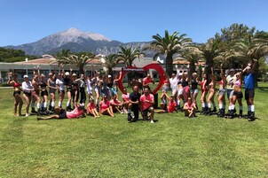 Un grupo de personas posando en un área con césped, con una montaña al fondo y un letrero rojo en forma de corazón que dice 'clubmarcopolo'.
