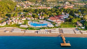 Vista aérea de un complejo turístico frente al mar, con piscina, playa y montañas al fondo.