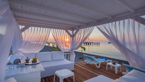 Un salón junto a la playa con muebles blancos, una mesa preparada para comer y vistas al atardecer sobre el agua.