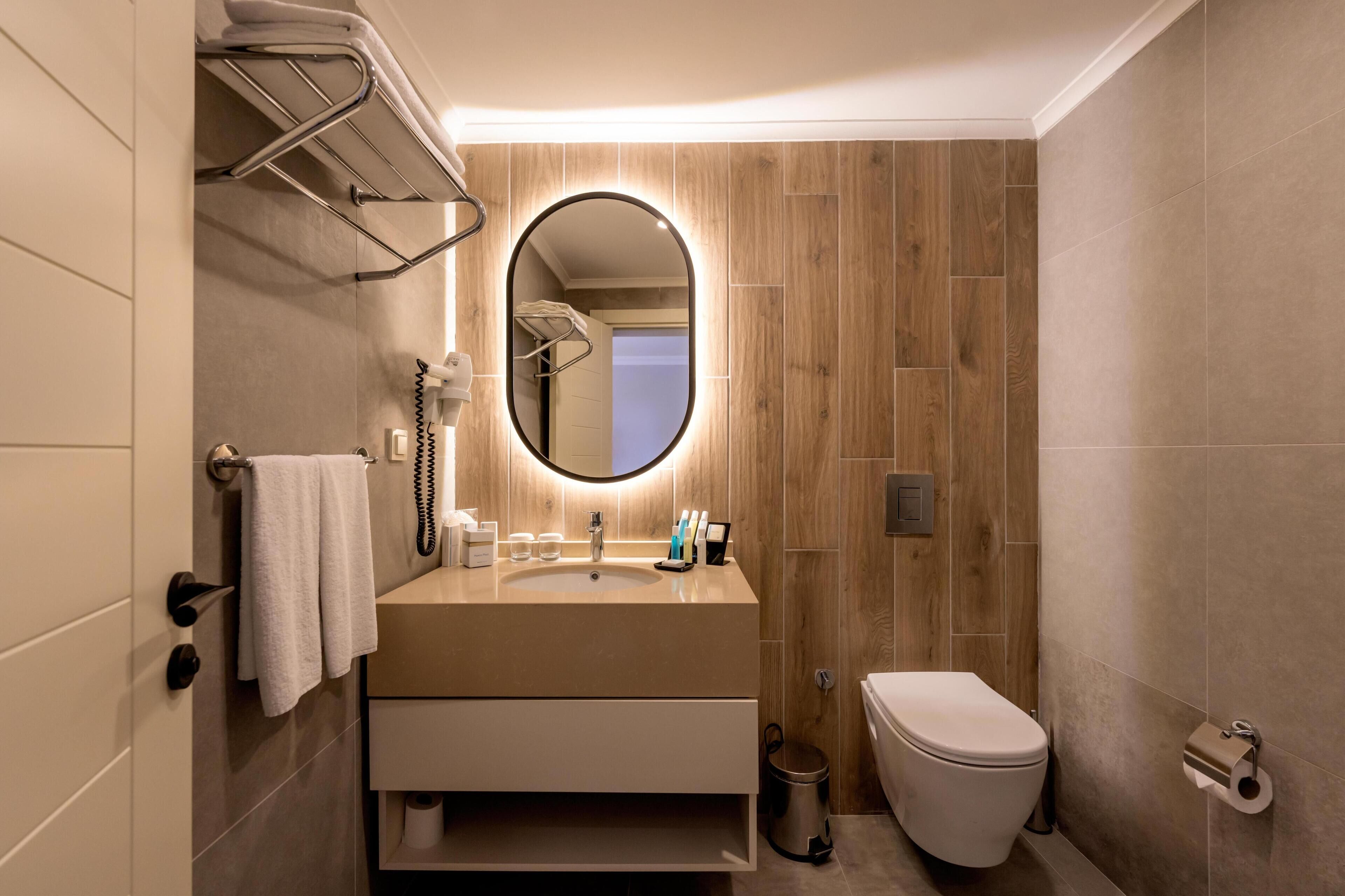 Un bagno moderno con parete in legno, uno specchio rotondo, un lavandino con articoli da toilette, un WC e un porta asciugamani.