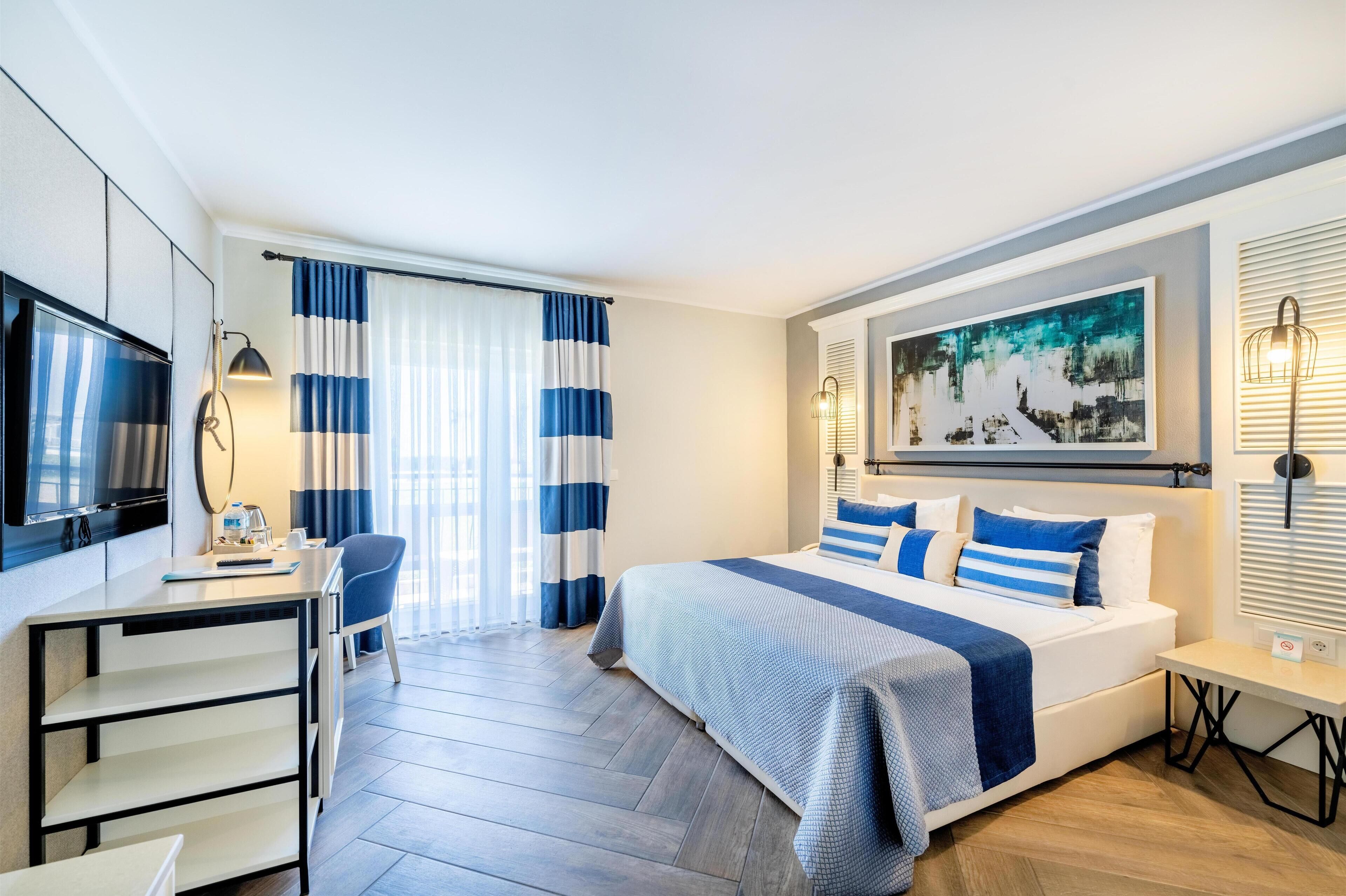A modern hotel room with a bed, a desk, a chair, a TV, and a painting on the wall.