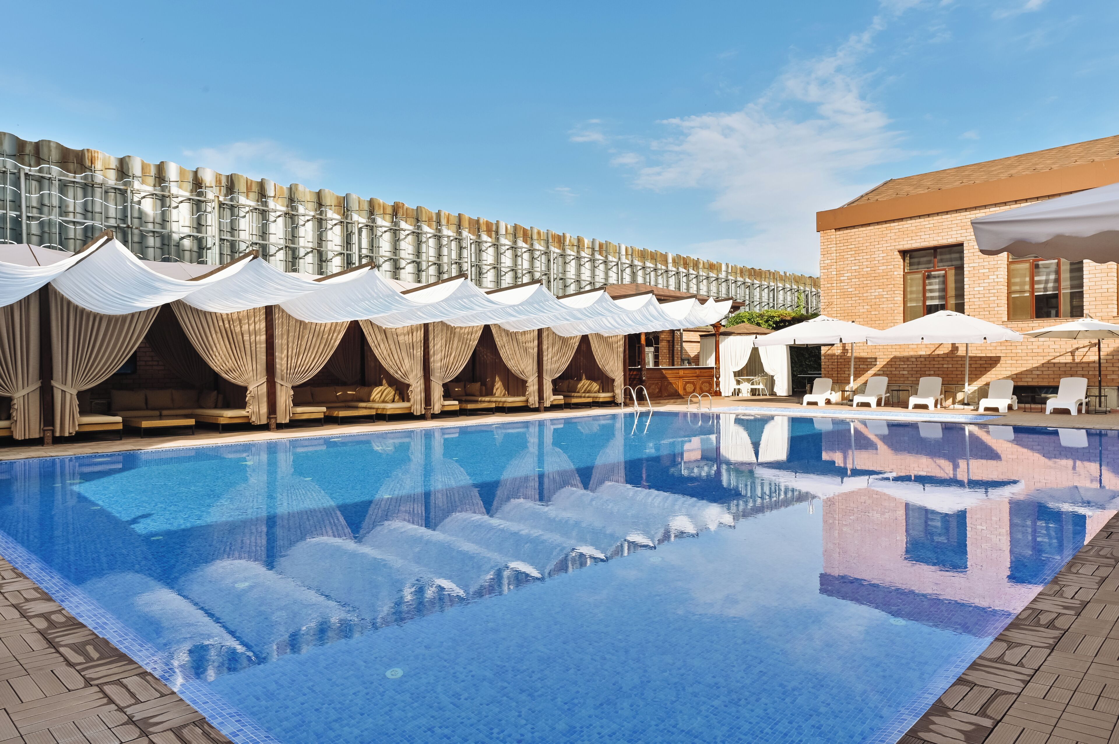A swimming pool with lounge chairs and umbrellas, a brick building, and a fence with draped fabric.