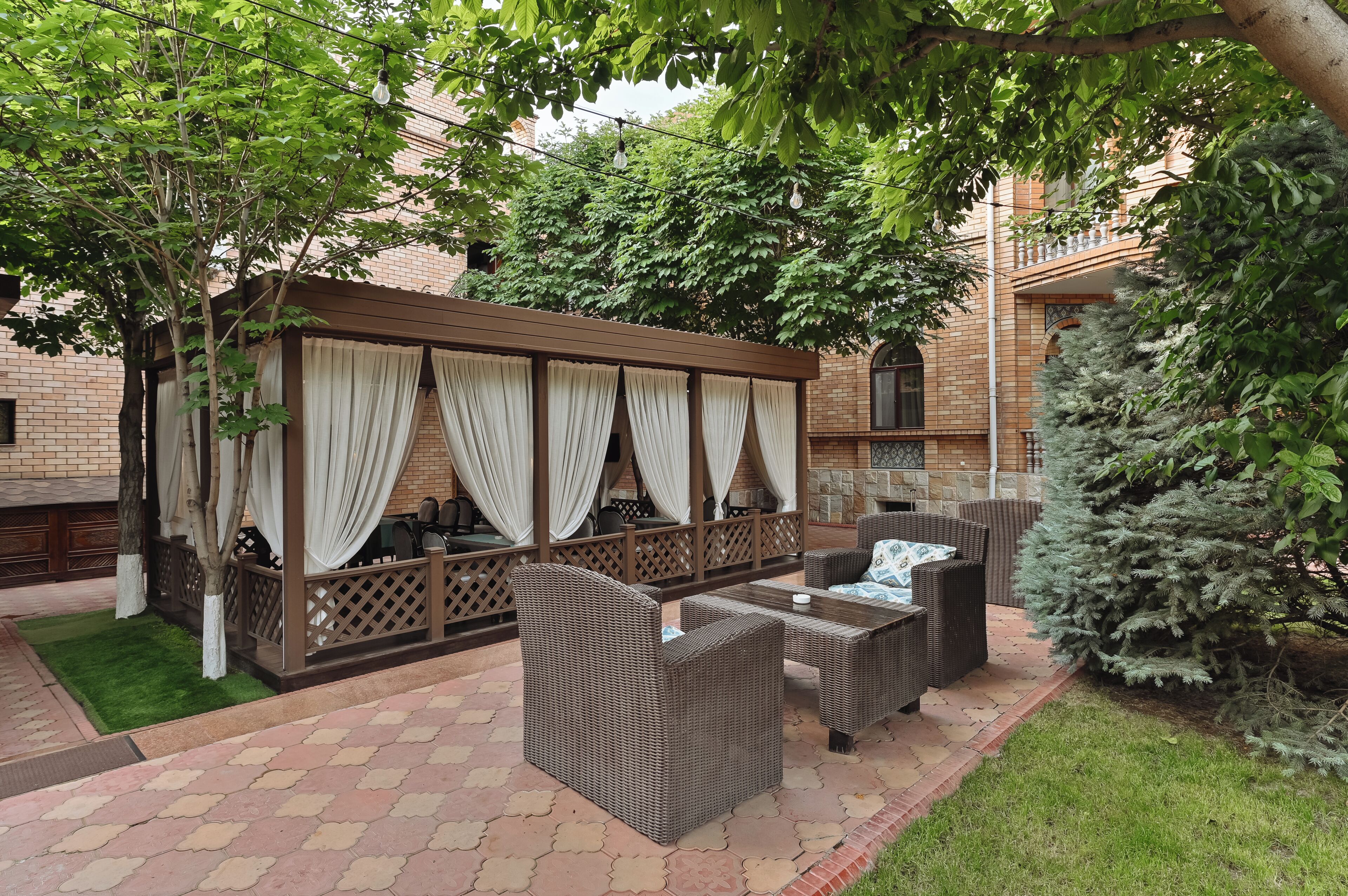 An outdoor seating area with wicker furniture, a pergola with draped curtains, and a brick building in the background.