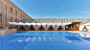 Una piscina con un edificio de ladrillo al fondo, una zona de estar cubierta y un bar.