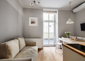 A modern living room with a sofa, a dining table with chairs, a kitchen area with a stove, and a large window providing natural light.