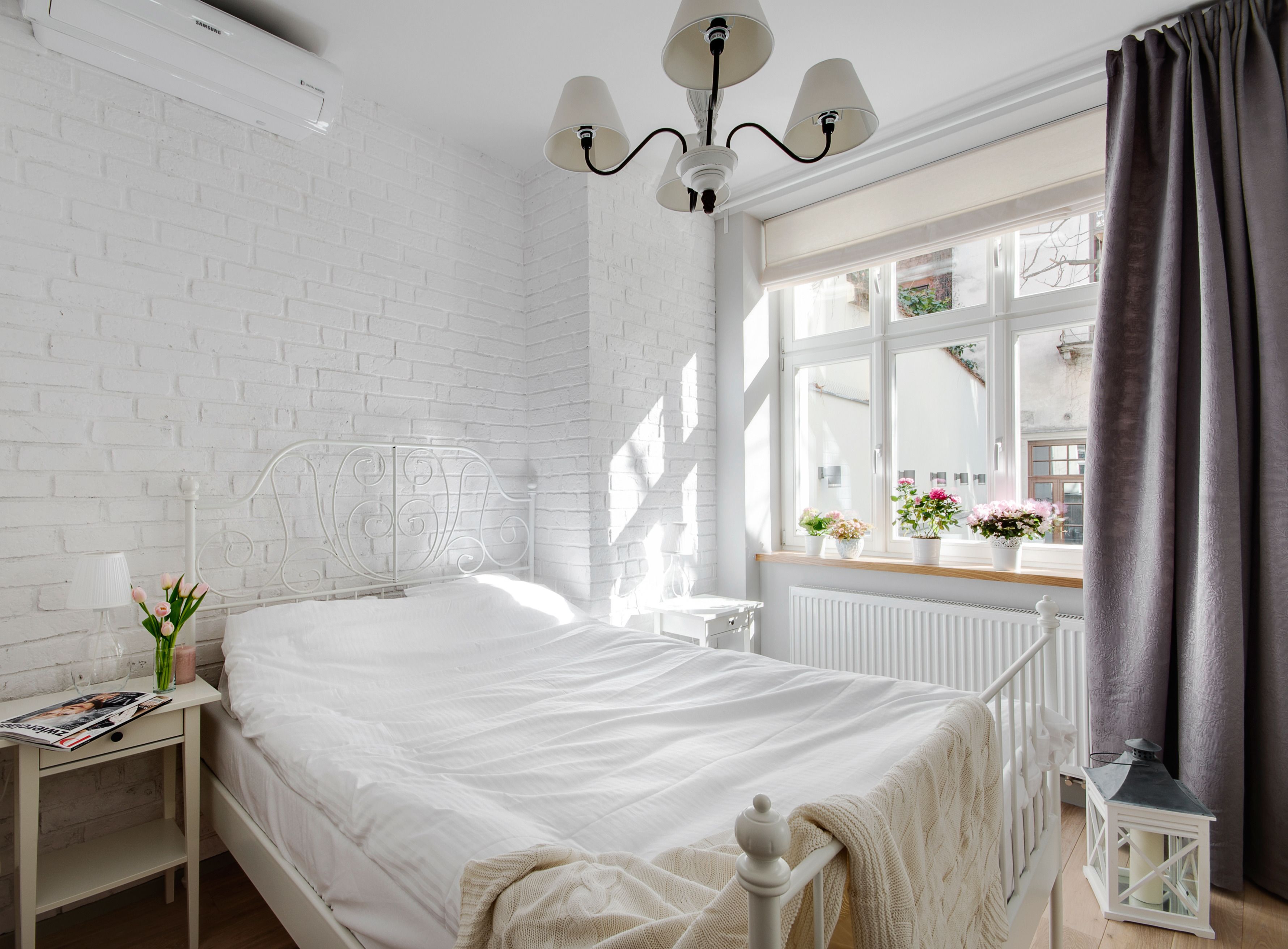 A bedroom with a white brick wall, a white bed, a nightstand with a lamp, a window with curtains, and a radiator.