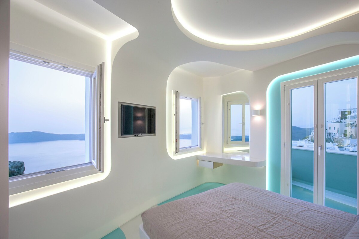 A modern bedroom with a bed, a TV, and large windows offering a view of the sea.