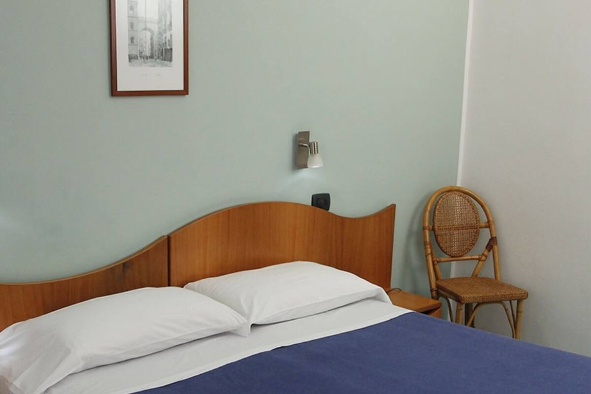 A bed with white and blue bedding, a wooden headboard, a wicker chair, a wall-mounted lamp, and a framed picture.