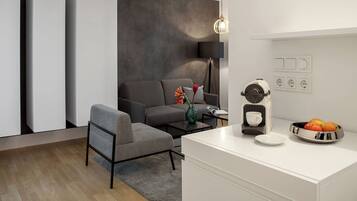 A modern living room with a grey sofa, a small armchair, a coffee machine, and a fruit bowl on the counter.